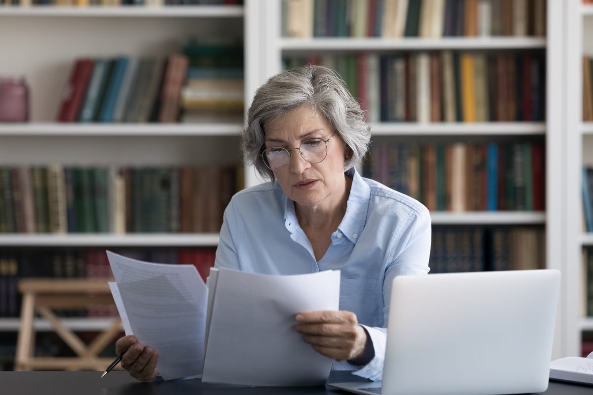 Serious thoughtful middle-aged woman in glasses looks worried read news in formal document sit at workplace desk with wireless computer